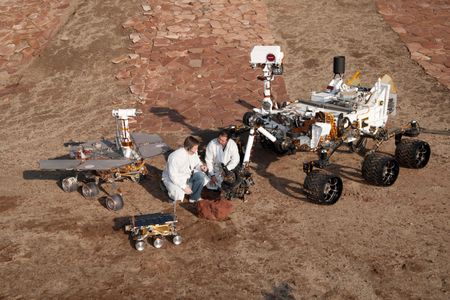 Scientists crouch with mock-ups of three generations of Mars rovers. Curiosity is the big one. Opportunity and Spirit were based on the medium-sized one on the left. The small one is front was the Sojourner rover.