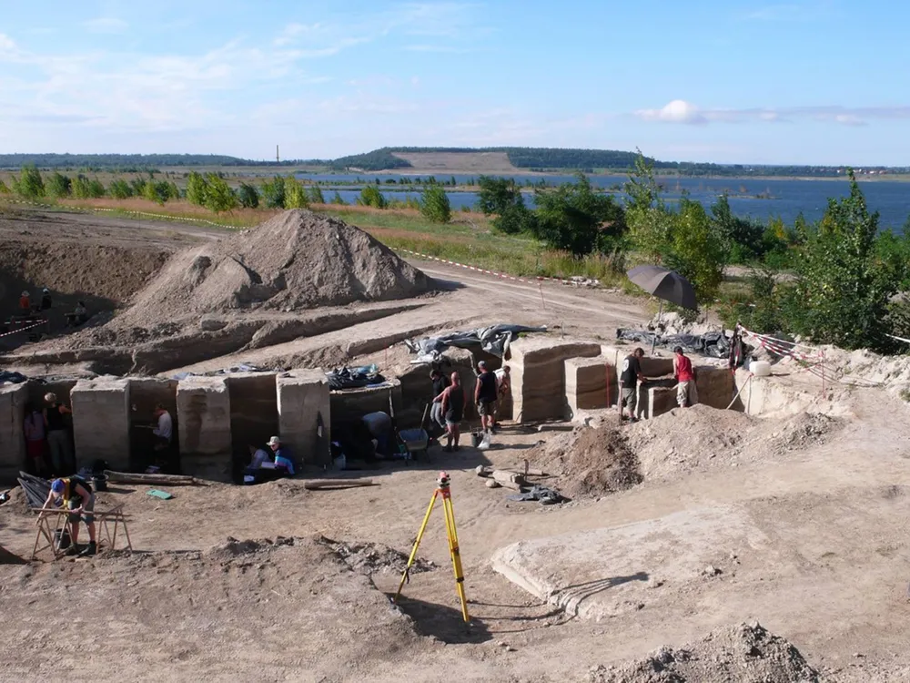 An image of archaeologists excavating a site. They stand in a pit near a mound of dirt. The surrounding area has lakes and trees.