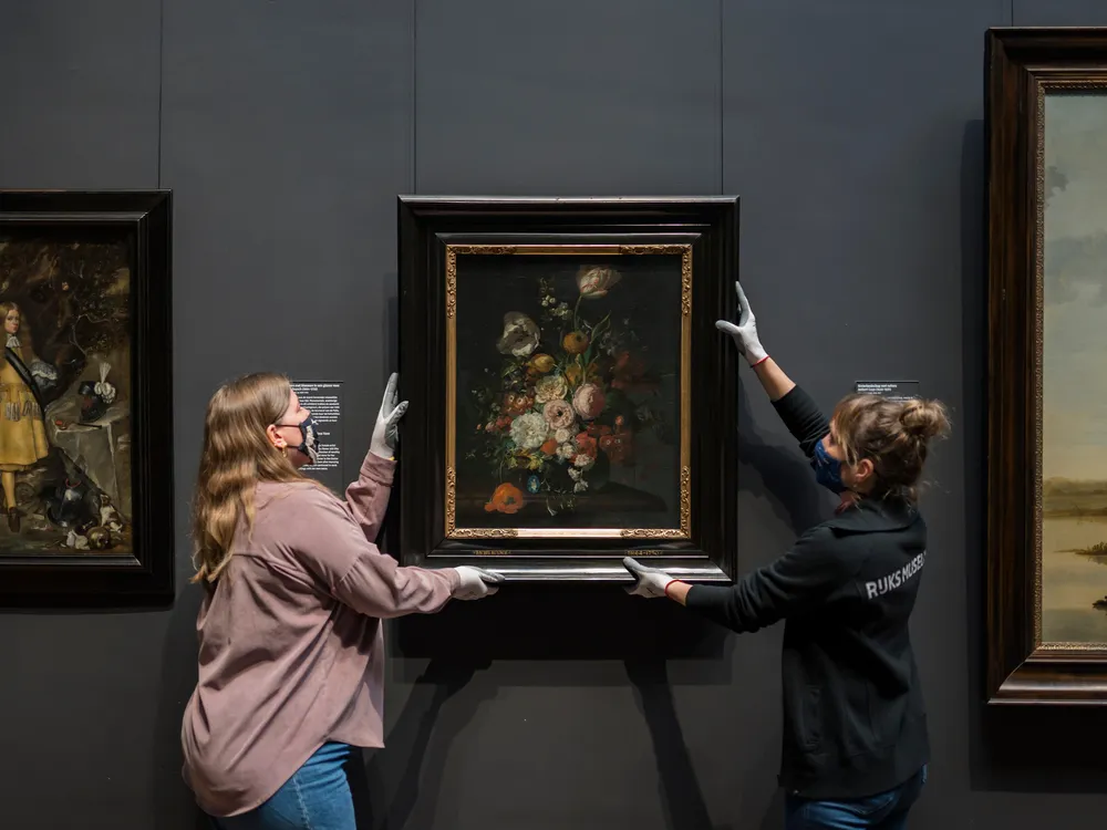 Two people in gloves and masks hold either side of the painting, which depicts a lush bouquet, and hangs it on the dark gallery wall
