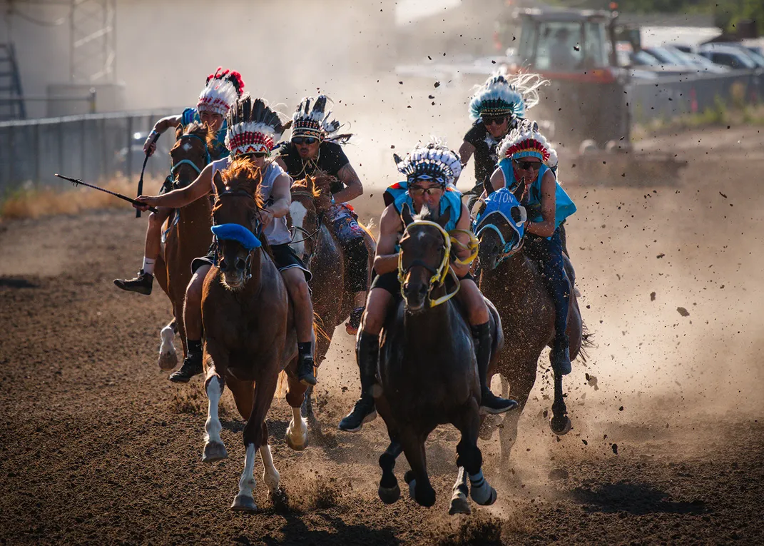 Several people in Native regalia, including feather headdresses, race on horseback down a dirt track, kicking up a cloud of dust.