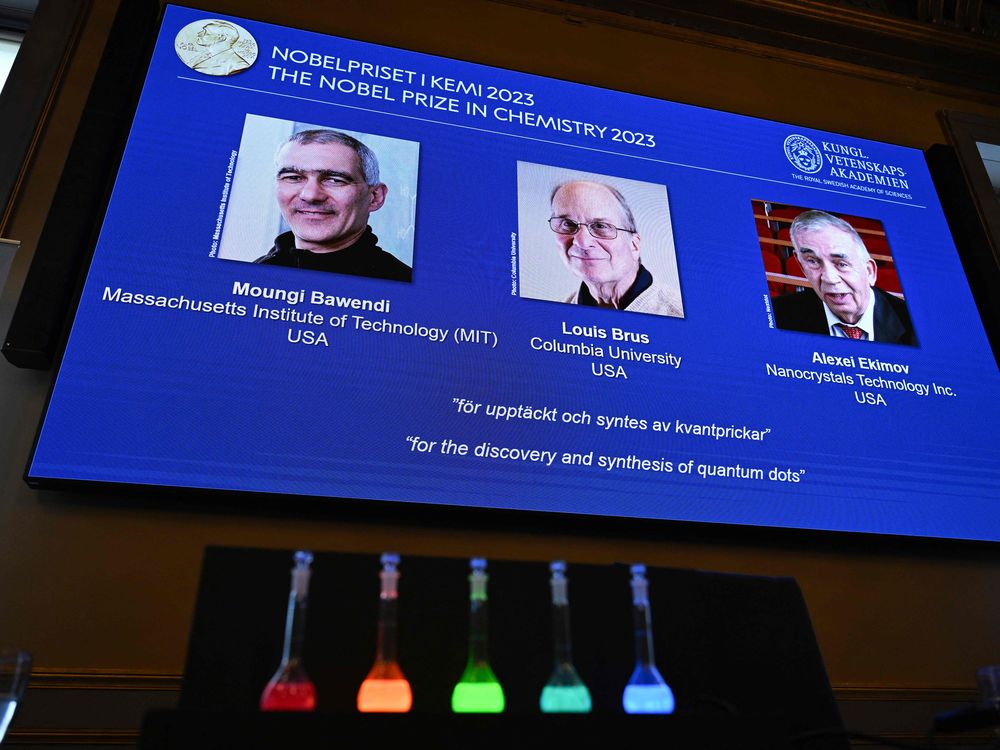 the winners of the chemistry nobel prize, Moungi Bawendi, Louis Brus and Alexei Ekimov, appear on a tv screen with five vials of glowing colors in front
