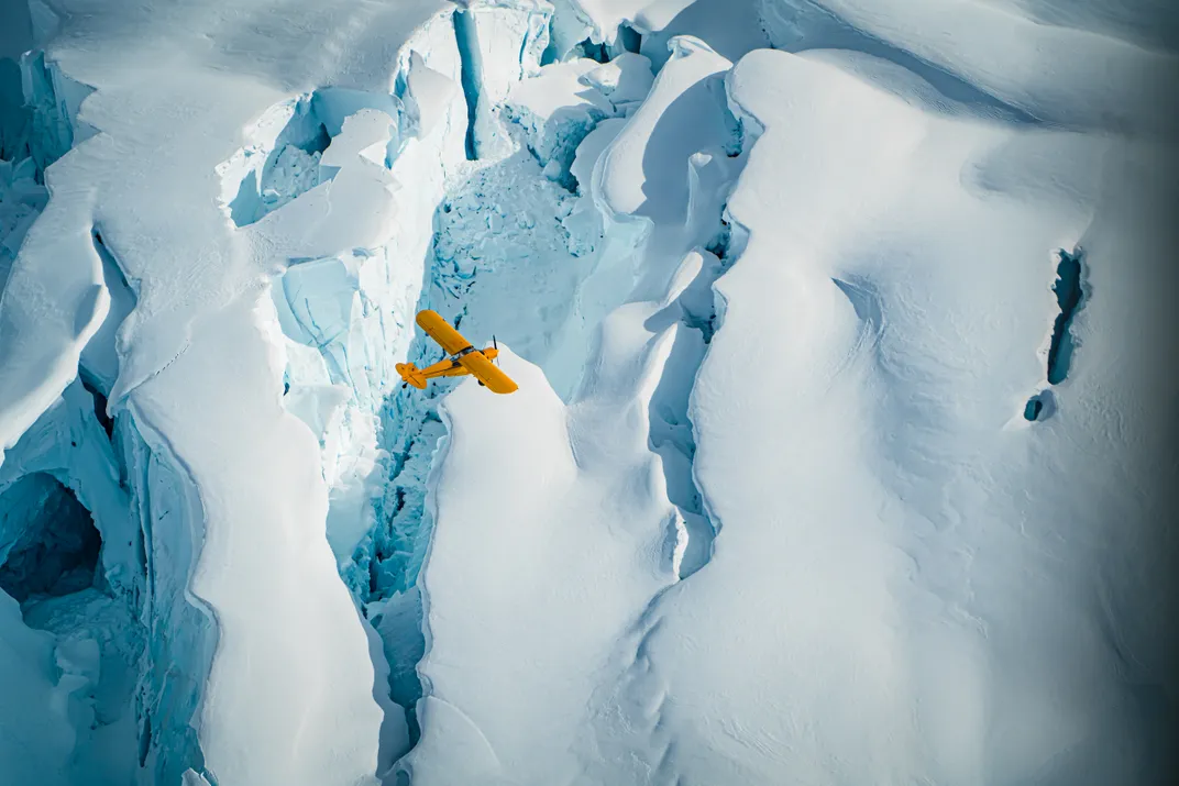 The bright yellow of a biplane stands in stark contrast to snow-white glaciers as it flies above.