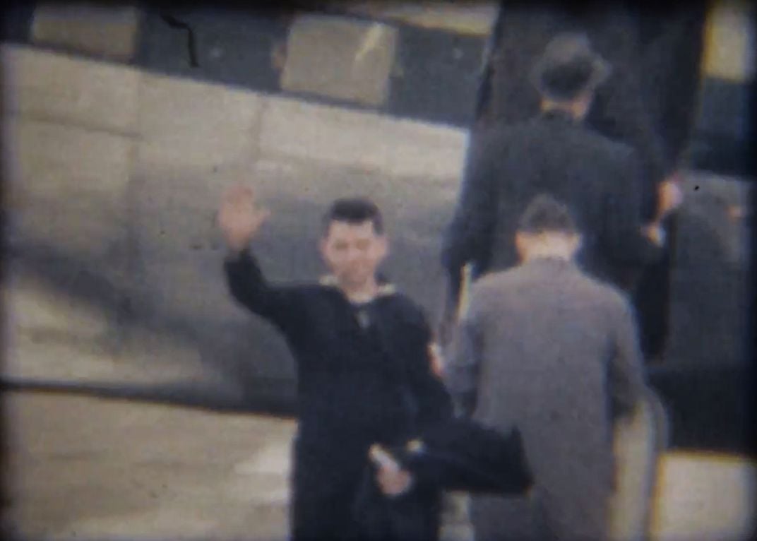 Old film still of a young man in Navy uniform, waving before boarding a plane.