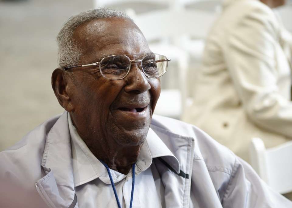A close-up of Brooks, a black man with glasses and gray hair, smiling and wearing a white collared shirt and overcoat; he is sitting down and uses a wheelchair (not pictured)