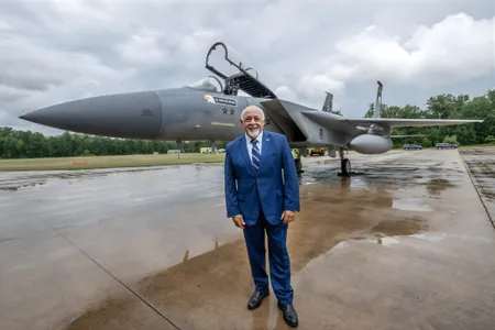 A 65-year-old man dressed in a blue suit poses in front of a military jet on a rain-soaked ramp.