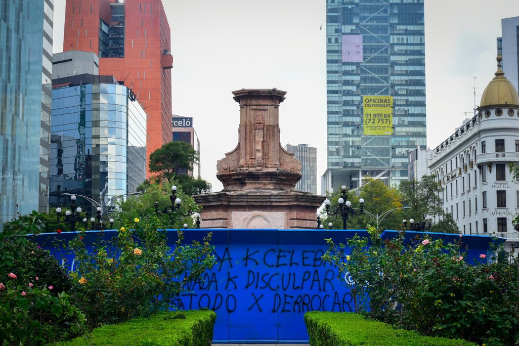 An empty pedestal stands, surrounded by tall buildings on all sides and a bright blue metal fence covered in graffiti
