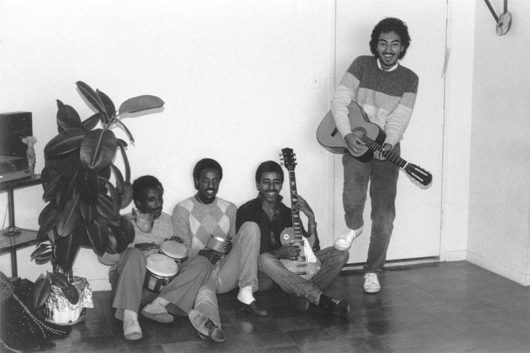 Four men pose, three sitting on the ground with a house plant towering over them. They all hold instruments: bongos, a shaker, electric and acoustic guitars. Black-and-white photo.
