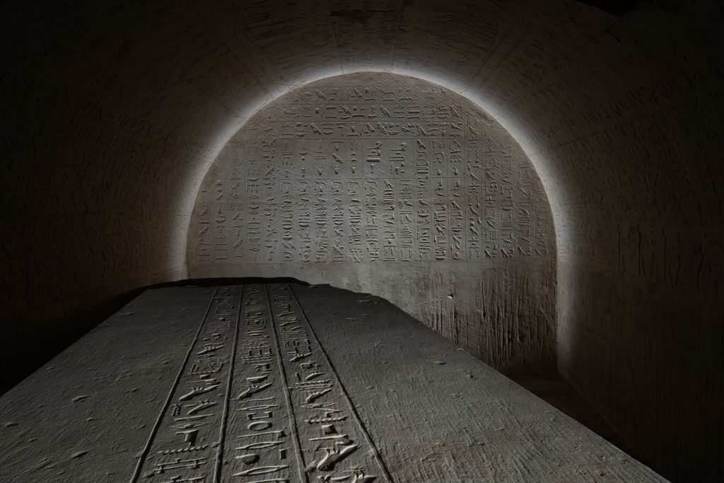 the top of a sarcophagus covered in writing with a glyph-covered wall behind it lit in an arch