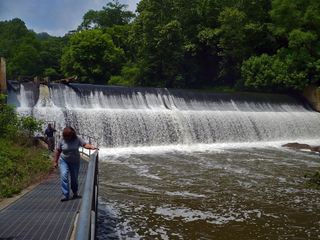 The Bloede Dam creates a smooth waterfall sloping downward in the river. On the left, two people walk on a boardwalk above the water near the dam.