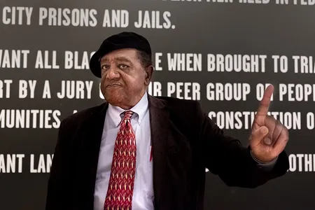 Founder of the Black Panther Party Bobby Seale stands next to a wall graphic of the Party's Ten-Point Program inside the Oakland Museum of California's exhibition, "All Power to the People: Black Panthers at 50."