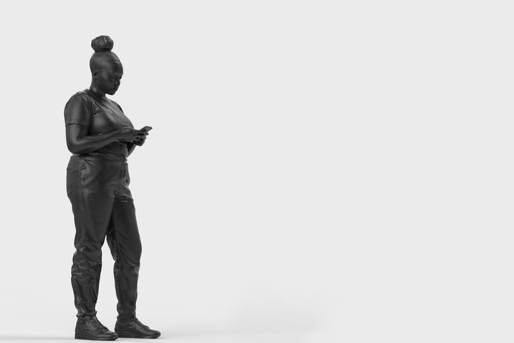 A bronze statue of a black woman surrounded by a plain white background. The woman is standing casually and looking down at her mobile phone, which she holds with both hands.