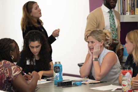 Teachers converse at a table, one woman with blond hair has her hands to her face