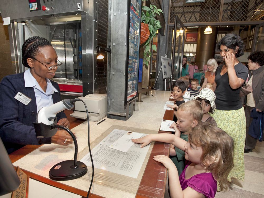 A group of students wait in line at a Post Office. Each student has a stamped envelope they are handing to the postal worker behind the counter.