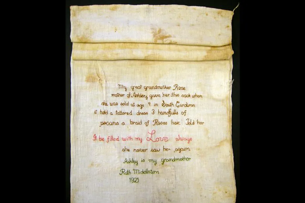 A yellowed, stained sack made from pale white cotton, with its top two thirds roll and its lower segment extended to reveal an embroidered message, stitched in elegant looping script 