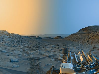 A stunning panorama of the Martian desert with a twilight sky. The landscape appears light rust and dull blue and is covered with boulders. A hill can be seen on the right.