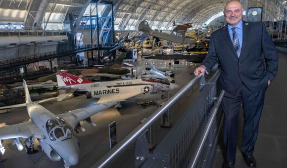 A white man in his mid-50s stands on a mezzanine overlooking several military jet airplanes parked on the concrete floor of a large hangar. The man is smiling and wearing a dark gray suit, blue shirt, and tie.