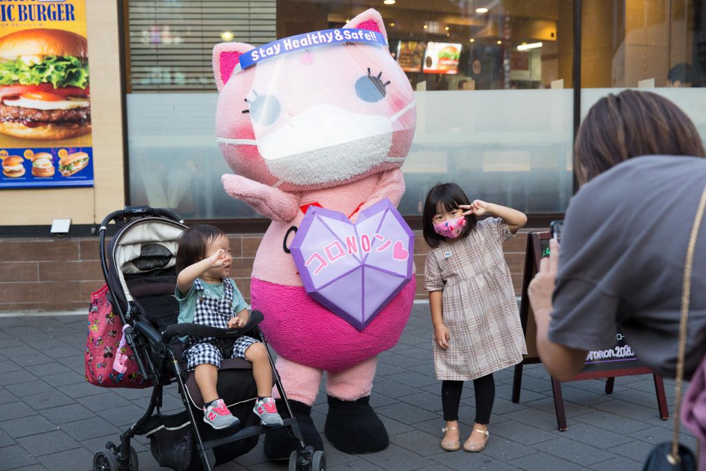 A photo of Japan's Covid-19 awareness Mascot, Koronon, a pink cat with a mask and a purple shield, poses for a photo with two young children. 