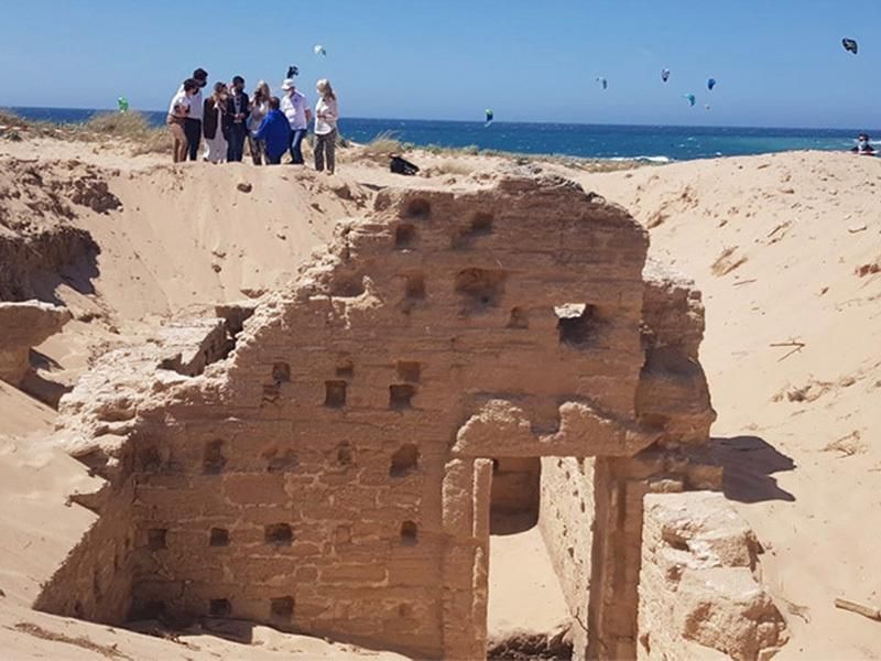 A group of people stand in front of a blue sky and blue sea while looking at a structure, with many small holes like windows and tall walls