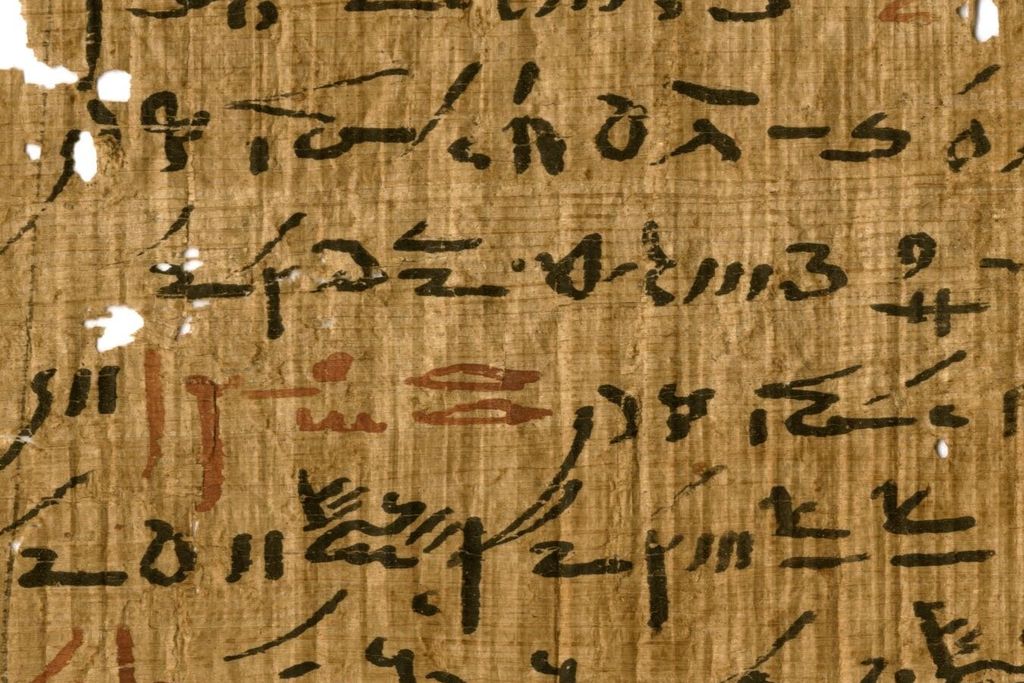A yellow-orange papyrus sheet, with some small holes, with elongated handwritten black hieroglyphic script, with small sections every few lines written in faded red ink