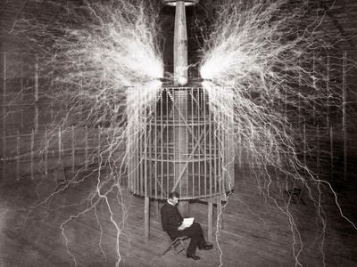 Both genius and impresario, Serbian-American inventor Nikola Tesla reads in his remote Colorado Springs laboratory in 1899 next to a magnifying transmitter that generates millions of volts of electricity. While far too dangerous to sit near—the image is a double exposure—his gigantic Tesla coil created the first human-made lightning.