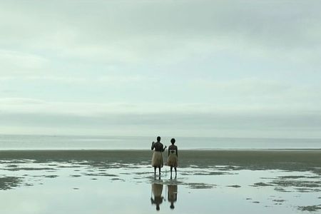 Two people in grass skirts stand on the shore, facing the ocean under an overcast sky.