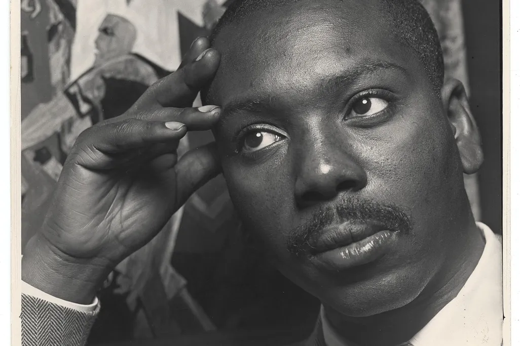A black and white image of Lawrence, a black man with a mustache, looking contemplative with a suit and tie and resting his head against his hand, in front of one of his works of art