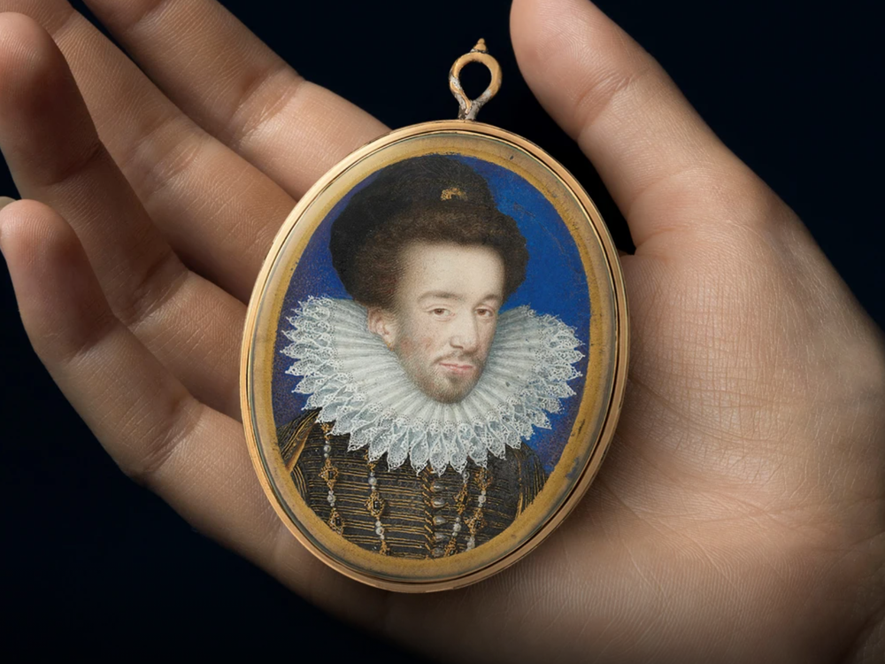 A white hand holds a small portrait in its palm; the oval frame is gilded and holds a portrait of a white man framed by blue, a starched ruffle around his neck and elaborate jewels
