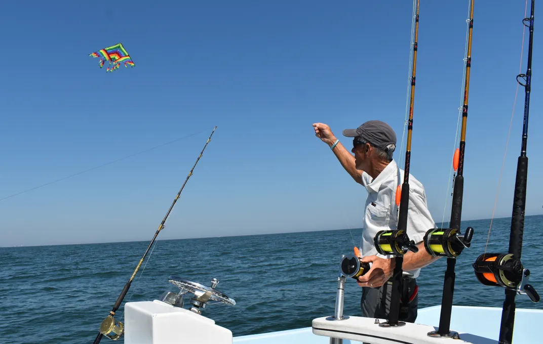 a main raises a colorful kite with streamers off a boat, surrounded by fishing rods