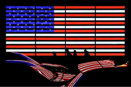 An illuminated American flag display casts its colors onto passing cars, merging national symbolism with New York City&rsquo;s constant flow of people and light.