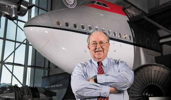 A white man in his early 70s stands inside a museum gallery. He is wearing glasses and smiling. Behind him, is the nose and forward fuselage of a Boeing 747 airplane. The 747 is painted in a red, gray, and white paint scheme.