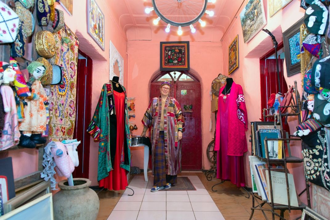 Lola Sayfi stands in Human House, surrounded by clothing, paintings, and crafts, and she smiles at the camera.