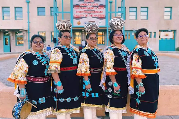 Five women in Zuni Pueblo garments stand in line, three with decorated ceramic pots balanced on their heads, looking at the camera.