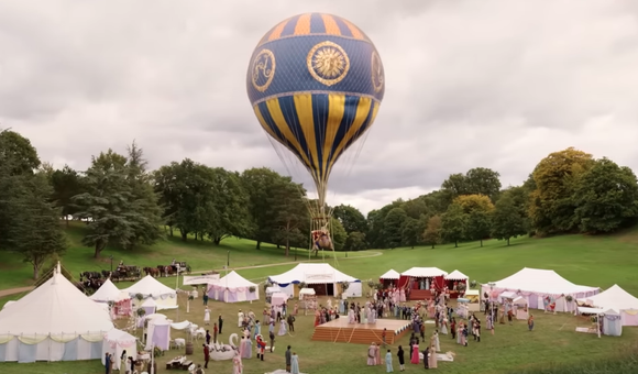 A vibrant hydrogen balloon with intricate blue and gold designs floats above a bustling outdoor event set against a cloudy sky. Below, several white tents are set up in a grassy park, with groups of people mingling and walking around.