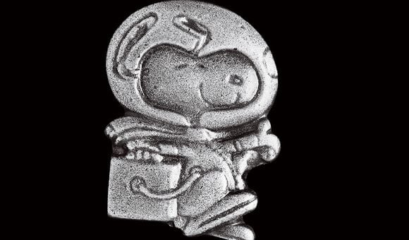 A small silver pin shows a smiling Snoopy, a cartoon dog, wearing a spacesuit with a large clear glass helmet, performing his iconic happy beagle dance.