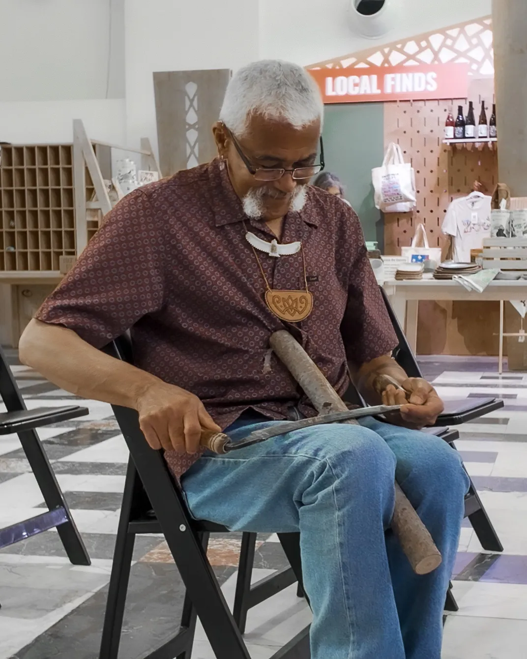 A man with white hair and moustache and glasses, seated, holds a cylindrical wooden flute between his knees, shaping it with a metal tool.