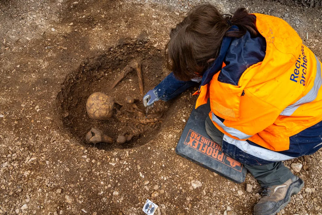 A person wearing a bright orange coat leaning over a skeleton in a hole in the ground
