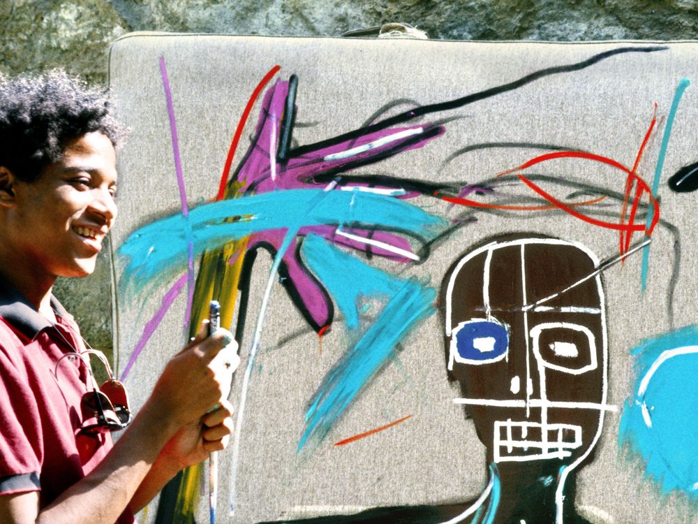 Jean-Michel Basquiat, smiling, stands in front of one of his works in progress. It's sunny outside, and he wears a red button down shirt