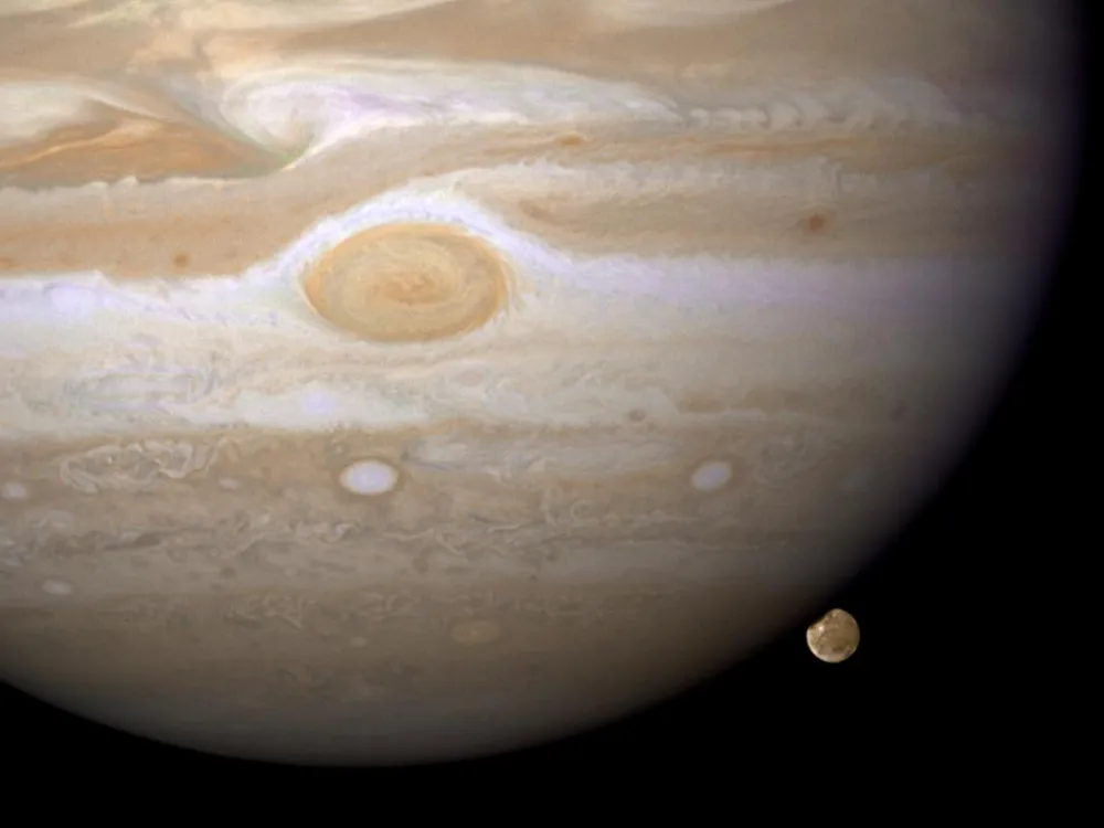 Spanning most of the image is the planet Jupiter, its red spot centrally featured. In the lower right, its largest moon Ganymede peaks from behind the planet.