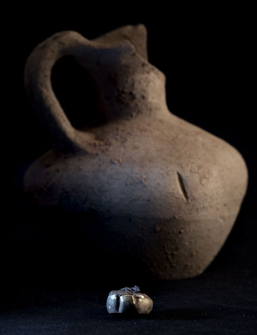 A gold earring in the foreground, with a brown pot in the background.