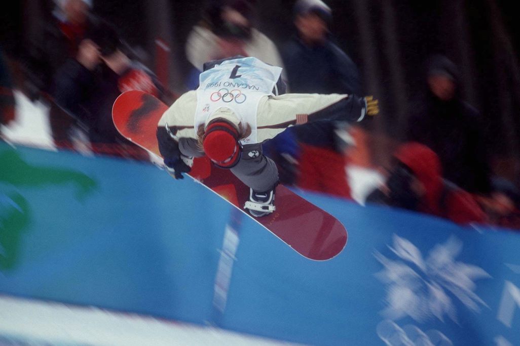 snowboarder performs trick at 1998 olympics, reaching down to touch the board