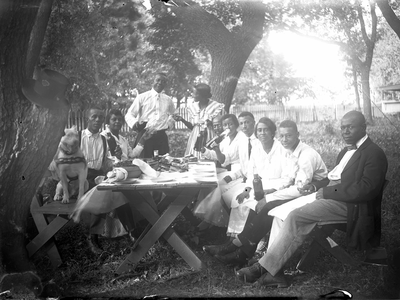 Photograph of ten people and a dog at a picnic table, 1919–1925