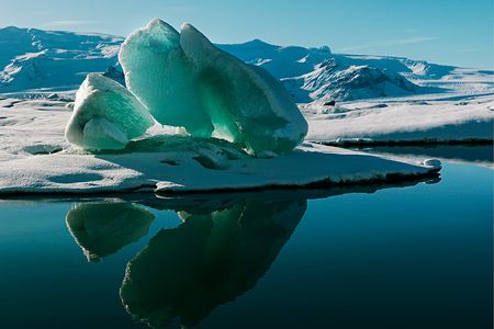 Lake Jökulsárlón shimmers with the reflection of a magnificent iceberg. This lake, located at the edge of Vatnajökull, Iceland’s largest ice cap, formed slowly when part of the glacier began to recede in the 1920s. The glacier continues to calve (split), releasing more icebergs into the expanding lake. 