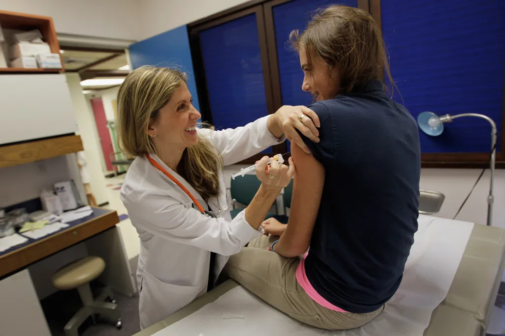 An image of a pediatrician giving an HPV vaccination to a young 13 year old girl in a doctor's office. Both subjects are smiling at each other.