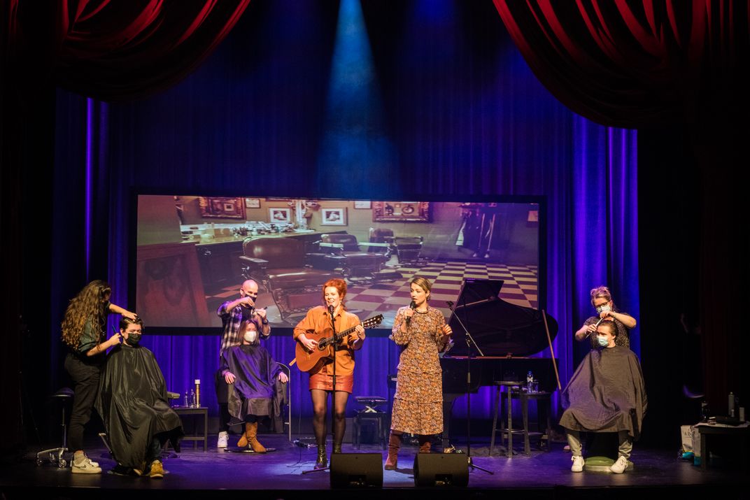 Actors perform on stage while two people receive haircuts, one on each side of the theatre stage