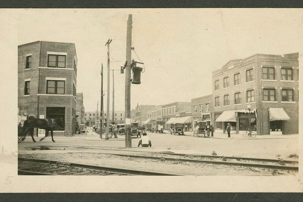 A yellow-toned black and white image of a cross-street, with a horse and carriage on the left, a Black person sitting in center frame, and many businesses and storefronts lining a city street