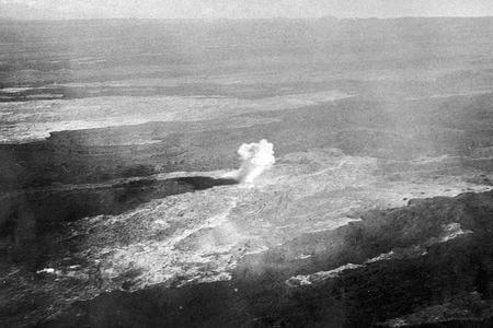 An old photo, tinted brown, depicts a flat barren landscape with a single pillar of dense white smoke at the center of the photo.