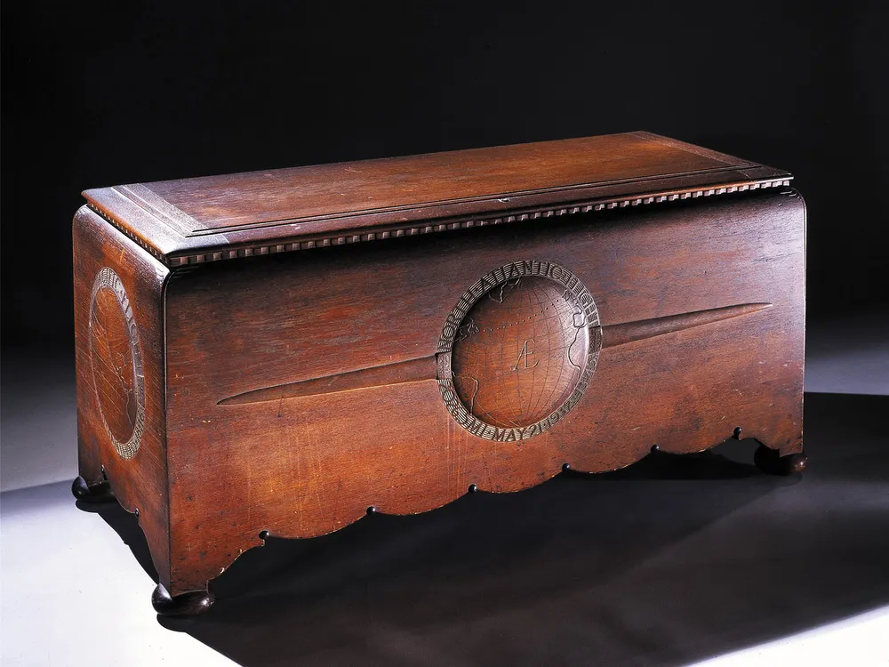 A large hand-carved teakwood chest with scalloped edges and four round feet. The chest appears to be stained a rich reddish brown. The front panel on the chest has a bas-relief globe pattern flanked by a pair of airplane wings.