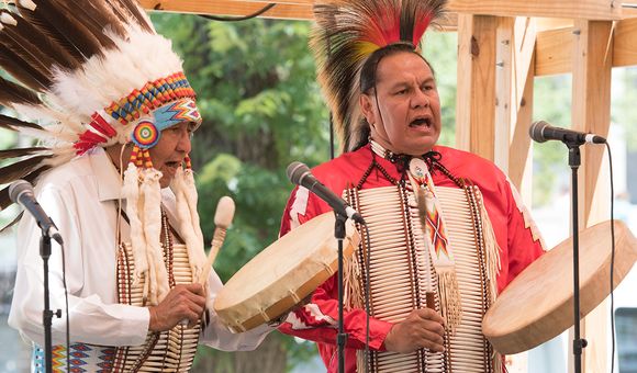 Two men wearing traditional feathered headdresses stand in front of two microphones, singing and playing hand drums.