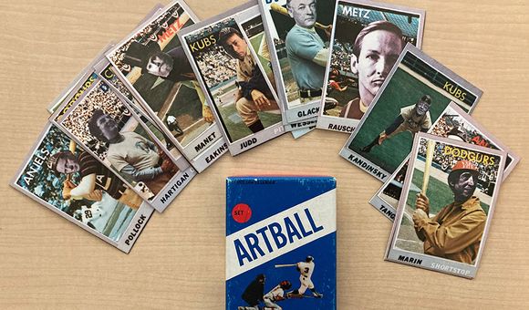 Baseball cards with faces of artists superimposed on them fanned out on a desk above a blue box with text that reads ARTBALL, an image of a batter, catcher, and umpire, with a red circle that says set 1.