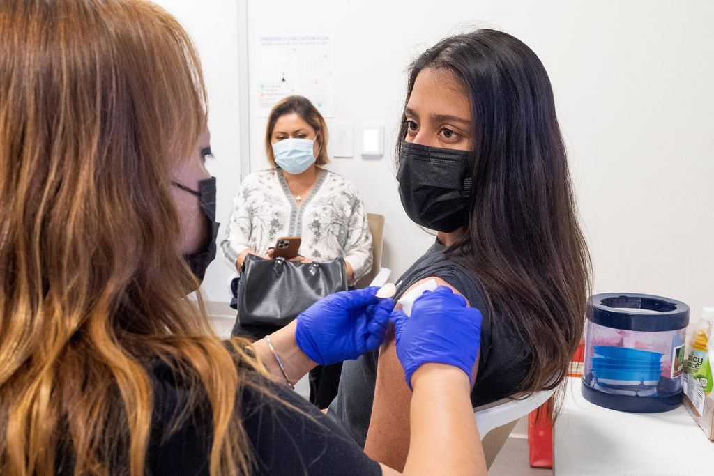 The back of a nurse's head is out of focus on the left side of the image. To the right, a teen girl sits with her sleeve rolled up facing the nurse as she places a bandage on the teen's shoulder.